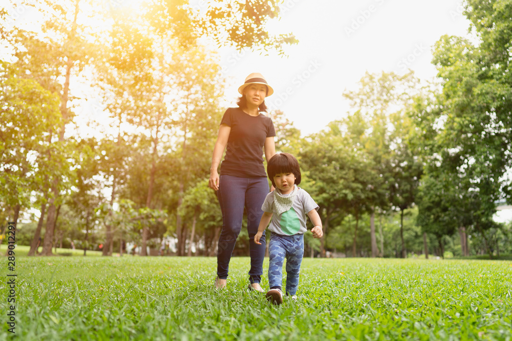 Fototapeta premium Cute 2 year old asian toddler girl exploring the park with excitement while the mother follow and walking behind protective in the natural warm sunlight tone, concept protective mom and child love.