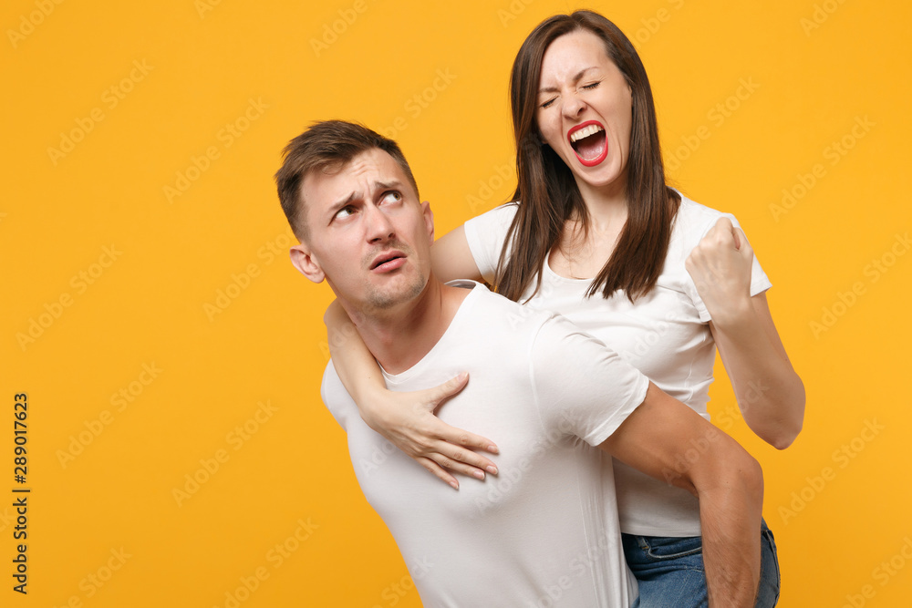 Young couple in white t-shirts posing isolated on yellow orange background. People lifestyle concept. Mock up copy space. Giving piggyback ride to joyful sitting on back, clenching fist like winner. Young couple in white t-shirts posing isolated on yellow orange background. People lifestyle concept. Mock up copy space. Giving piggyback ride to joyful sitting on back, clenching fist like winner.