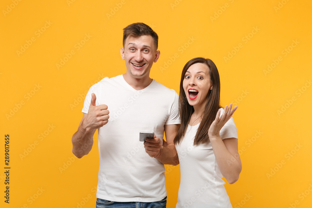 Cheerful young couple friends guy girl in white empty blank t-shirts posing isolated on yellow orange background. People lifestyle concept. Mock up copy space. Hold, using cellphone, showing thumb up. Cheerful young couple friends guy girl in white empty blank t-shirts posing isolated on yellow orange background. People lifestyle concept. Mock up copy space. Hold, using cellphone, showing thumb up.