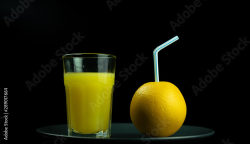 A glass of orange juice stands next to a yellow orange, which is inserted into the tube for drinks. Black background.