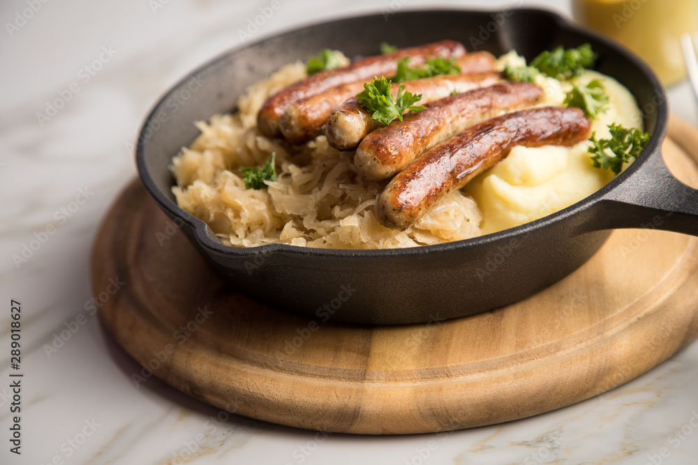 Kleine Bayrische Nürnberger Rostbratwürste mit Sauerkraut, Kartoffelpüree und Senf in rustikaler Gusseisenpfanne auf Holz Brett und Marmor Tisch Kleine Bayrische Nürnberger Rostbratwürste mit Sauerkraut, Kartoffelpüree und Senf in rustikaler Gusseisenpfanne auf Holz Brett und Marmor Tisch