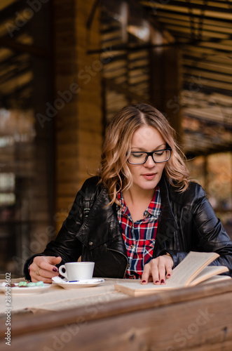 Attractive young woman sitting alone at the table in street cafe, drinking coffee or tea while reading book. Relaxing in cafe during free time