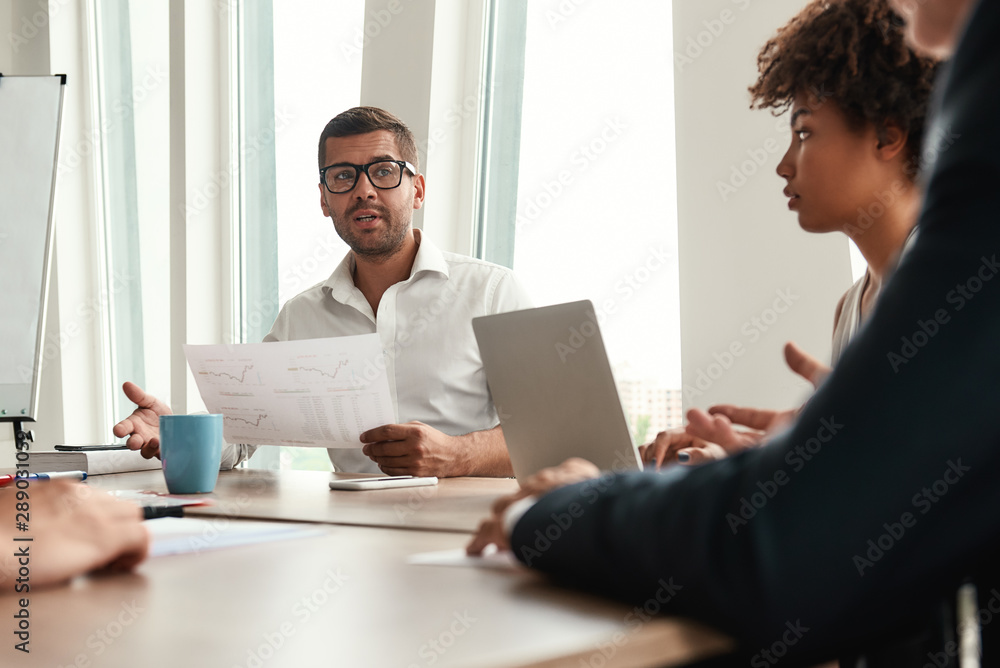 New business strategy. Young man in formal wear and eyeglasses holding financial reports and discussing it with team while sitting at the office table New business strategy. Young man in formal wear and eyeglasses holding financial reports and discussing it with team while sitting at the office table