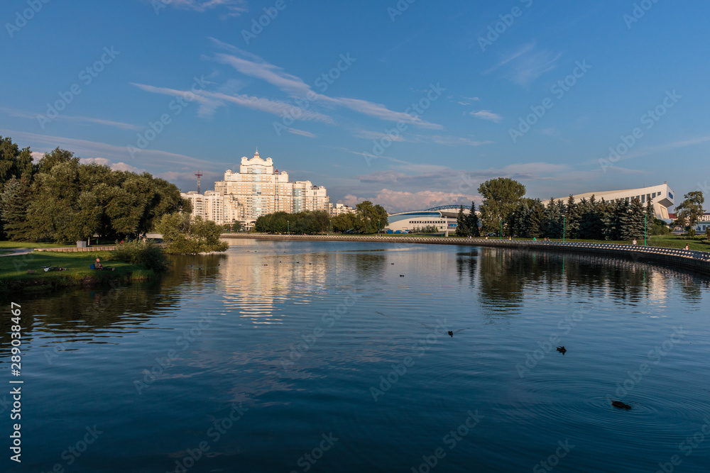 the river flows through it floating ducks on the background of a white building and green trees and above them the blue sky with Cirrus clouds