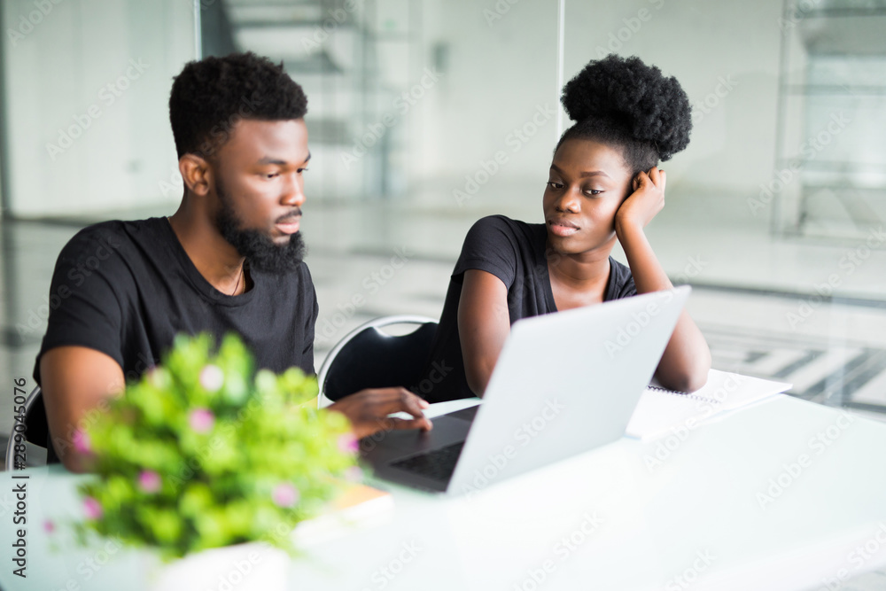Two young african coworkers working together in a modern office. African black business partners using laptop and discussing new startup project. Two young african coworkers working together in a modern office. African black business partners using laptop and discussing new startup project.