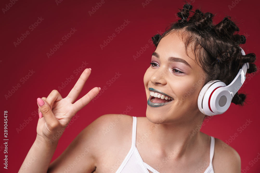 Naklejka premium Image of joyful punk girl with bizarre hairstyle and dark lipstick showing peace fingers while listening to music with headphones