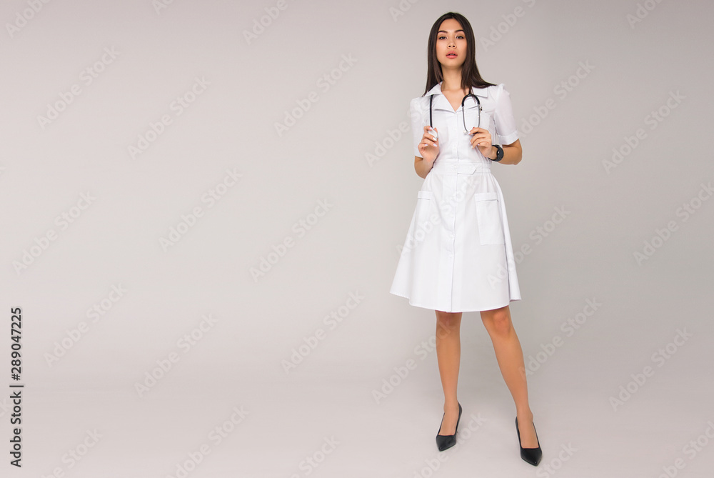 young stylish brunette nurse with long hair in white medical dress with stethoscope on her neck is standing on the white wall background. medical fashion concept. free space on left side young stylish brunette nurse with long hair in white medical dress with stethoscope on her neck is standing on the white wall background. medical fashion concept. free space on left side
