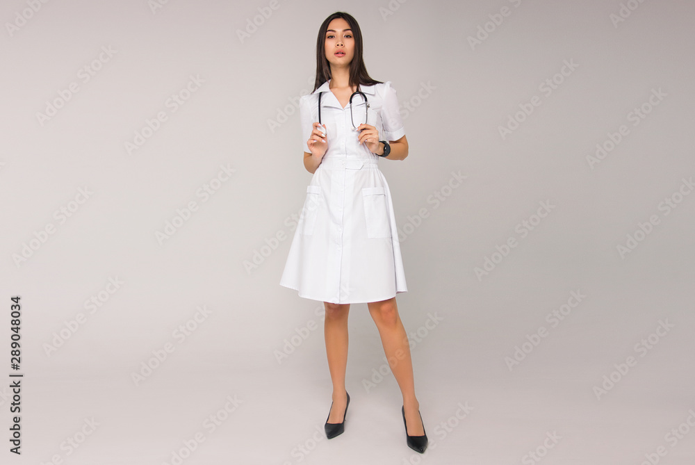young stylish brunette nurse with long hair in white medical dress with stethoscope on her neck is standing on the white wall background. medical fashion concept. free space