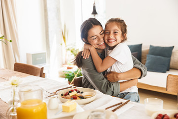 Image of happy family mother and little daughter hugging while having breakfast at home in morning