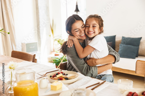Image of happy family mother and little daughter hugging while having breakfast at home in morning photo