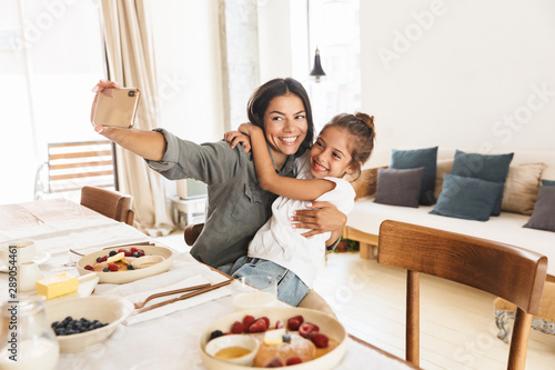 Image of beautiful family mother and little daughter taking selfie photo while having breakfast at home in morning