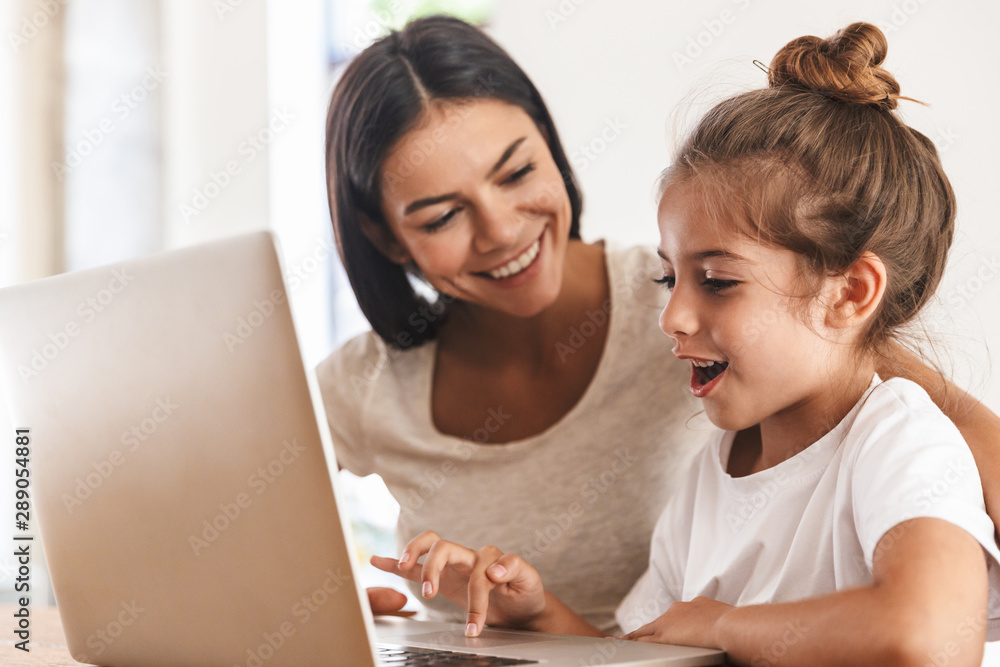 Image of joyful family woman and her little daughter smiling and using laptop computer together in apartment Image of joyful family woman and her little daughter smiling and using laptop computer together in apartment