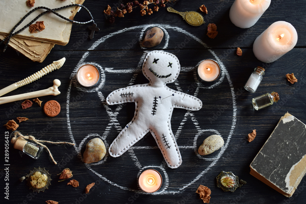 In Voodoo doll are needles pricked. Candles, pentagram, stones, love potion and old books on witch table. Occult, esoteric or divination concept. Mystic, Halloween and vintage background