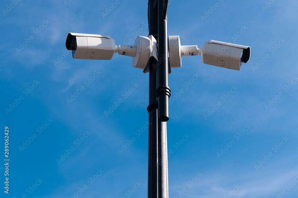Two white surveillance cameras on the metal lamp post against blue sky. Security of the whole territory. Security service is watching. Copy space. Two white surveillance cameras on the metal lamp post against blue sky. Security of the whole territory. Security service is watching. Copy space.