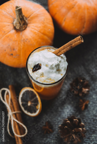 glass of fresh pumpkin juice with whipped cream and a stick of cinnamon, whole pumpkins, a slice of dried orange, cones on a knitted dark fabric