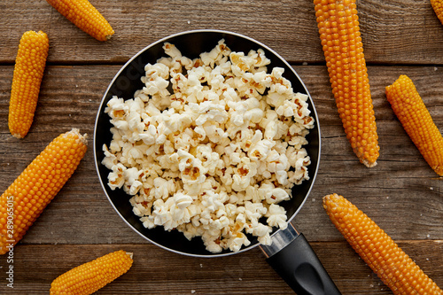 top view of tasty popcorn in frying pan near scattered corn on wooden background