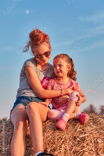 Sisters, teenage girl and her younger sister playing together on hay bale outdoors, spending summer vacation in the countryside. Candid people, real moments, authentic situations