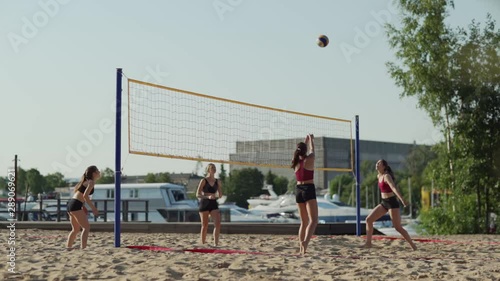 Side view of two teams of female athletes playing beach volleyball on sand court against each other, slow motion wide shot