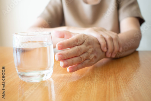 Elderly woman hands w/ tremor symptom reaching out for a glass of water on wood table. Cause of hands shaking include Parkinson's disease, stroke or brain injury. Mental health neurological disorder. photo