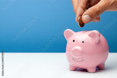 Pink piggy bank and coin in a male hand on a blue background. Copy space for text.