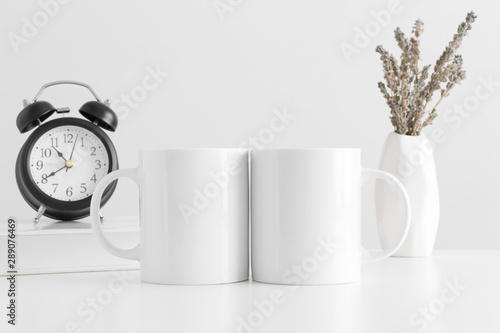 Two white mugs mockup with a lavender in a vase, book and a clock on a white table.