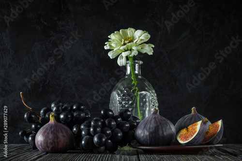 ripe whole and sliced figs with a bunch of black grapes and one flower in an empty decanter on old wooden boards against the background of a black textured concrete wall. elegant still life side view photo