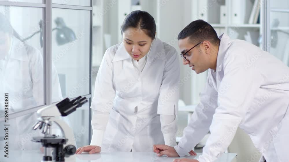 Medium shot of Asian woman and Arab man in white lab coats standing at desk with microscope beside them, discussing something and looking at invisible dimensional AR object. Suitable for AR, VR. Medium shot of Asian woman and Arab man in white lab coats standing at desk with microscope beside them, discussing something and looking at invisible dimensional AR object. Suitable for AR, VR.