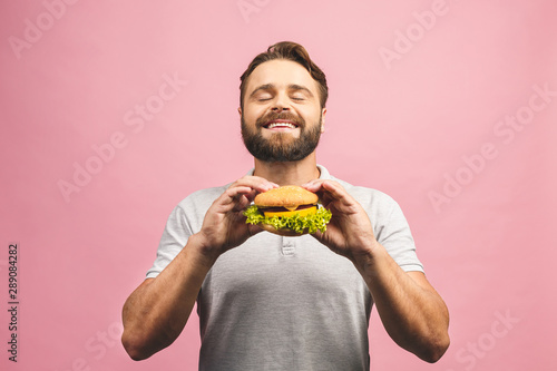 Young man holding a piece of hamburger. Bearded gyu eats fast food. Burger is not helpful food. Very hungry guy. Diet concept. Isolated over pink background. photo