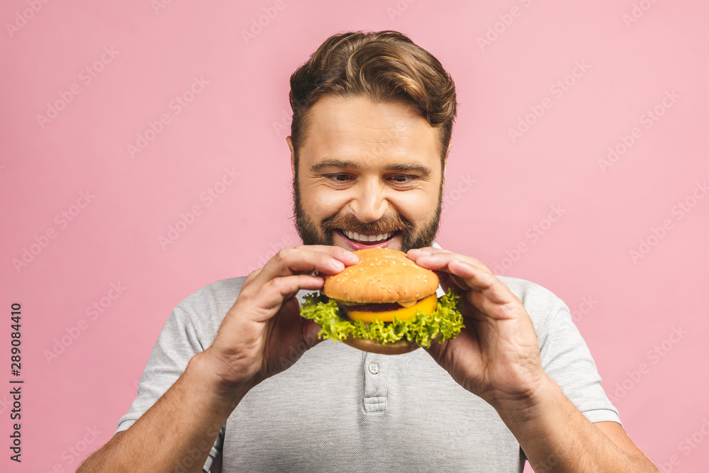 Young man holding a piece of hamburger. Bearded gyu eats fast food. Burger is not helpful food. Very hungry guy. Diet concept. Isolated over pink background. Young man holding a piece of hamburger. Bearded gyu eats fast food. Burger is not helpful food. Very hungry guy. Diet concept. Isolated over pink background.