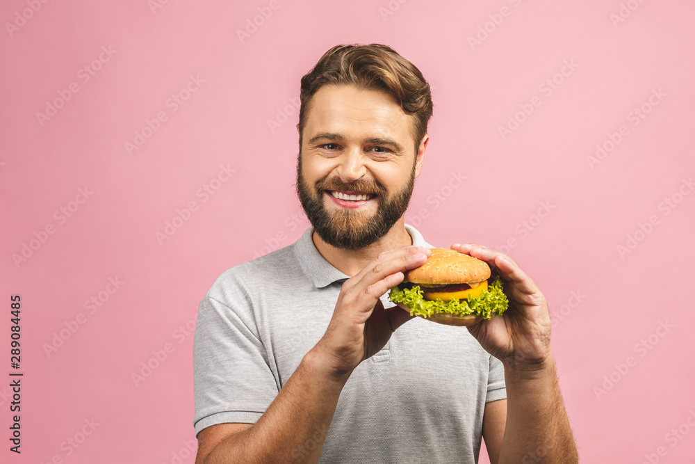Young man holding a piece of hamburger. Bearded gyu eats fast food. Burger is not helpful food. Very hungry guy. Diet concept. Isolated over pink background.