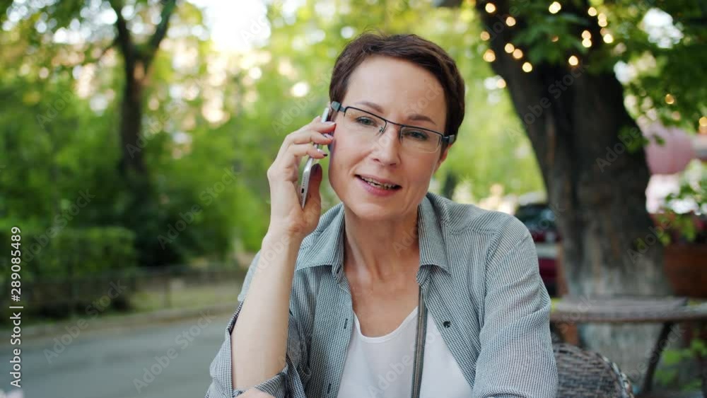 Mature woman in glasses is talking on mobile phone enjoying conversation smiling sitting in open air cafe outdoors. Conversation, emotions and people concept. Mature woman in glasses is talking on mobile phone enjoying conversation smiling sitting in open air cafe outdoors. Conversation, emotions and people concept.