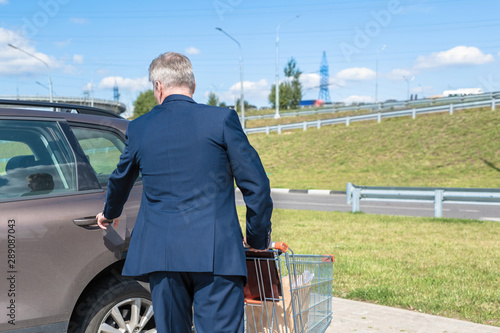 Elderly single man in a blue suit walks to his car with a supermarket trolley. Businessman and shopping