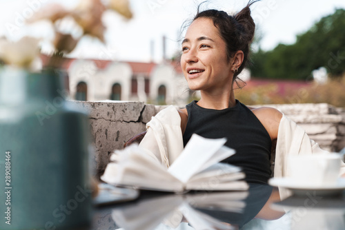 Portrait of pleased brunette woman reading book and drinking coffee while resting on terrace outdoors