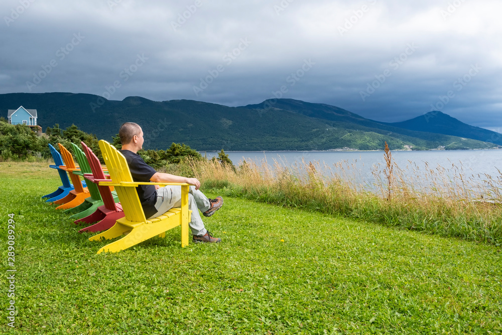 A Man Sitting in the First of a Row of Bright and Colorful Adirondack Chairs Facing Bonne Bay on a Cloudy Day in Newfoundland