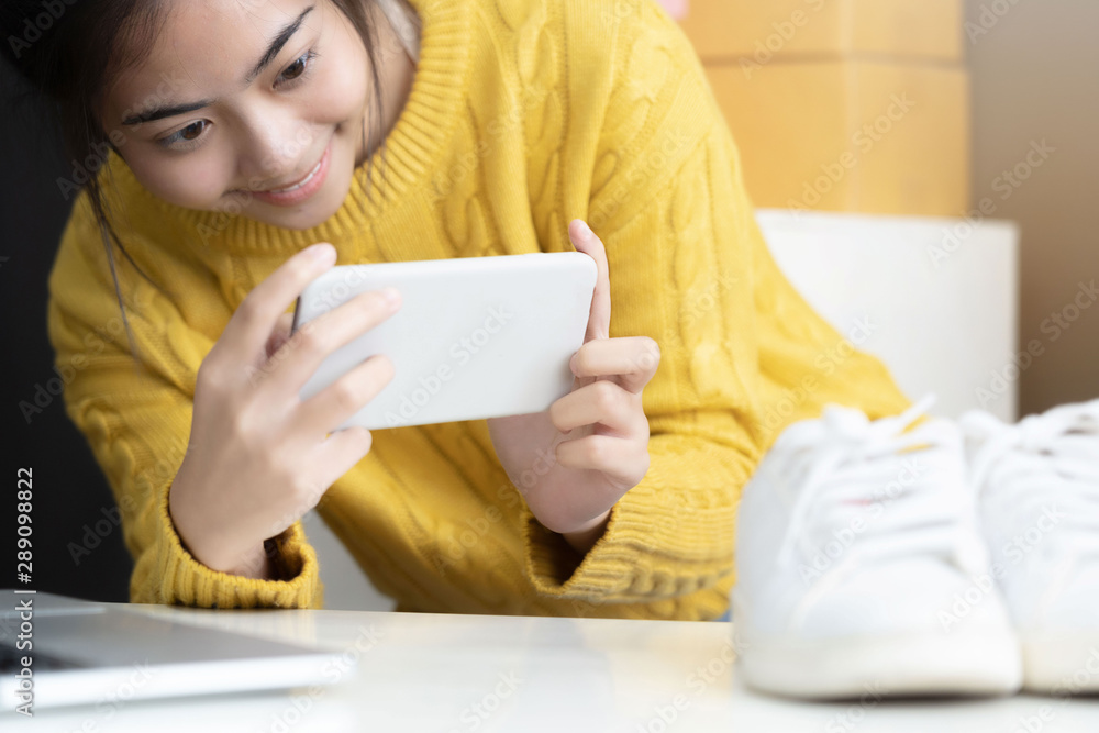 asian girl using smartphone to shoot white sneaker posted for sale on the internet.startup small business owner working with tablet at workplace.happy owner & seller check product order to customer asian girl using smartphone to shoot white sneaker posted for sale on the internet.startup small business owner working with tablet at workplace.happy owner & seller check product order to customer