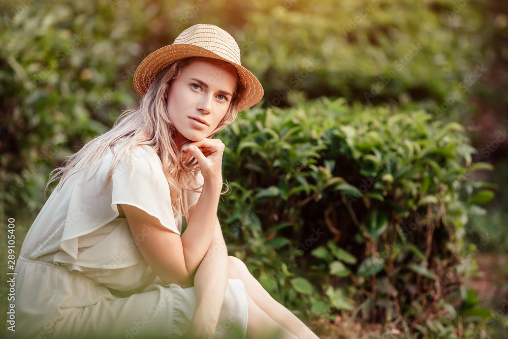 Portrait of a beautiful young blonde girl in a romantic white dress and straw hat posing on a background of green bushes on the sunny summer evening. Concept of environmental protection and ecology