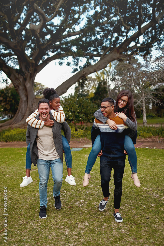 Fototapeta premium Happy young men carrying piggyback ride to their girlfriends in a park smiling - having fun outdoors