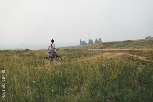 white european young woman in casual clothing going with bicycle on countryside road in hills, view from back in full body size, lifestyles stock photo image