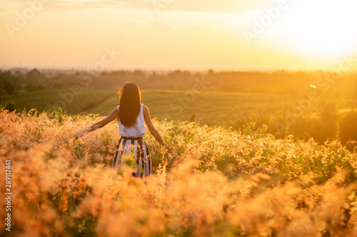 Trendy girl in stylish summer dress feeling free in the field with flowers in sunshine.