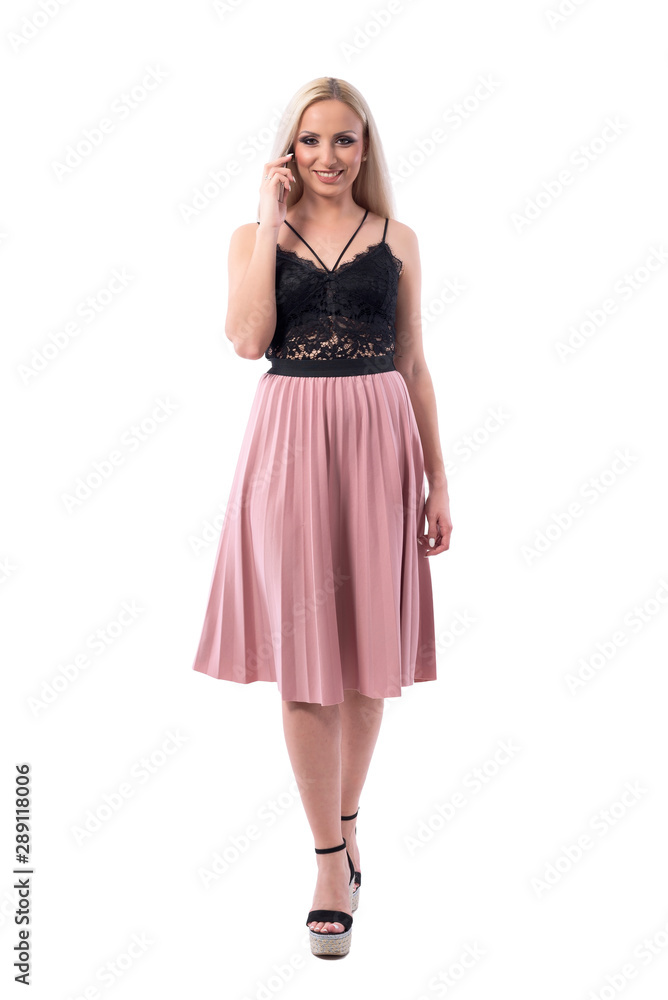 Confident happy young stylish blonde woman talking on the phone walking and looking at camera. Full body isolated on white background. Confident happy young stylish blonde woman talking on the phone walking and looking at camera. Full body isolated on white background.