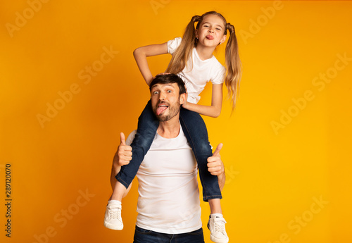 Man Carrying Daughter On Shoulders Gesturing Thumbs-Up Showing Tongues, Studio