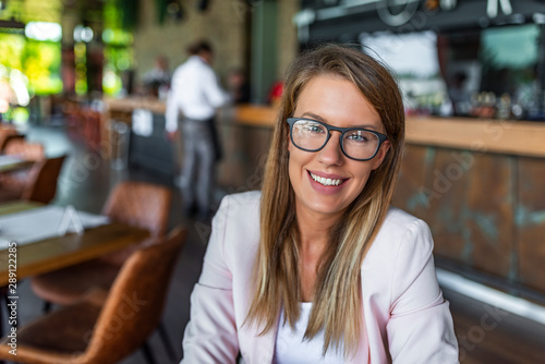 Portrait of smiling pretty young business woman in glasses sitting on workplace. Businesswoman look at you with confident smile on her face. Confident business expert.