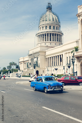 Photograph of a blue vintage car on the road in front of El Capitolio in Havana. In the background the dome of the building 