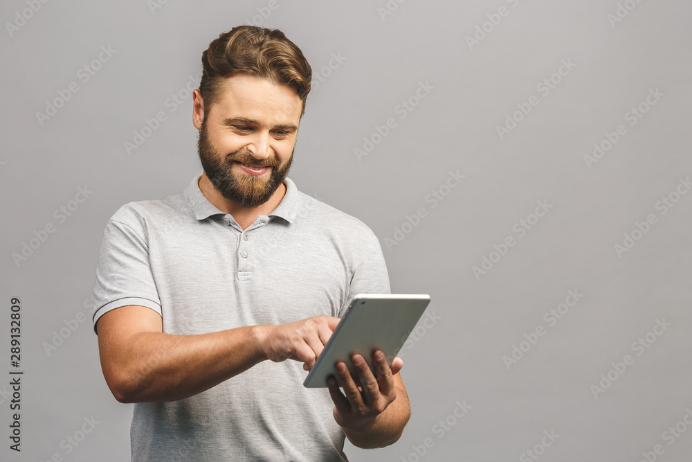 Happy bearded young man in casual standing and using tablet isolated over grey background. Happy bearded young man in casual standing and using tablet isolated over grey background.