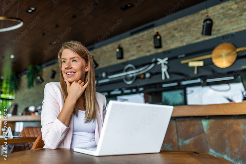 Beautiful business woman is using a laptop and smiling while working in coffee shop. Business woman sitting by the table with laptop. Beautiful young business woman in coffee shop Beautiful business woman is using a laptop and smiling while working in coffee shop. Business woman sitting by the table with laptop. Beautiful young business woman in coffee shop