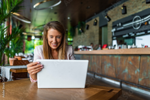 Beautiful business woman is using a laptop and smiling while working in coffee shop. Business woman sitting by the table with laptop. Beautiful young business woman in coffee shop