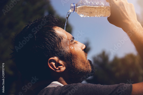Young man splashing and pouring fresh water from a bottle on his head to refresh against a blue sky background in a summer heat photo