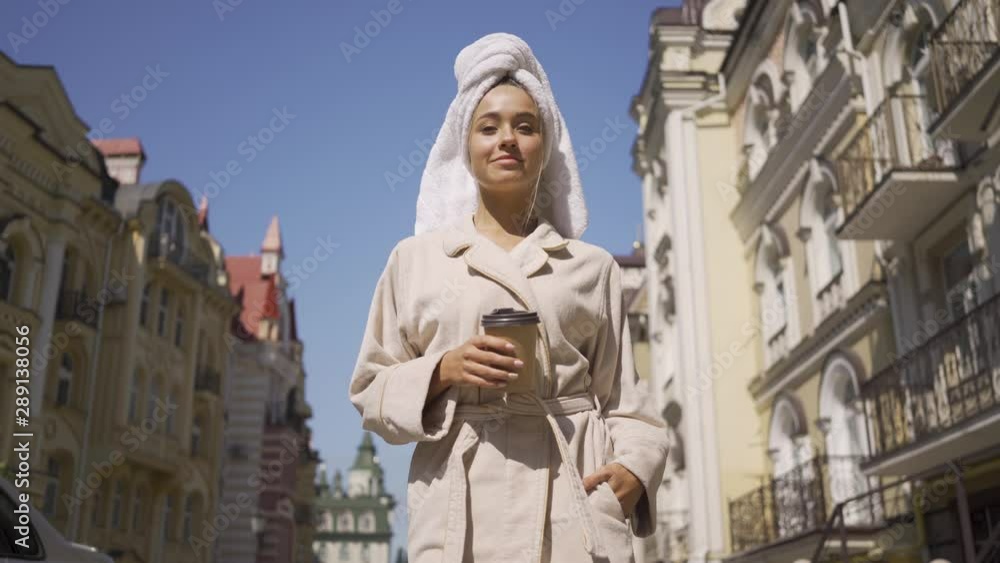 Pretty smiling young woman in bathrobe with towel on head walking down the street drinking coffee. Confident girl enjoying a beautiful day in the city Pretty smiling young woman in bathrobe with towel on head walking down the street drinking coffee. Confident girl enjoying a beautiful day in the city