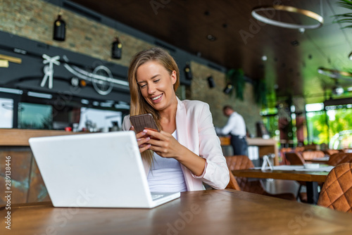 Digital displays surrounding us from everywhere. Sharing good business news. Attractive young woman talking on the mobile phone and smiling while sitting at coffee shop and looking at laptop