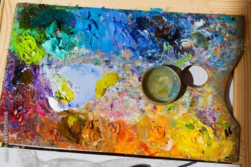 painter's wooden palette with mixed fresh bright oil paints, in disorder lays on wooden easel, summer plainair painting session, artistic flatlay in bright sunshine photo